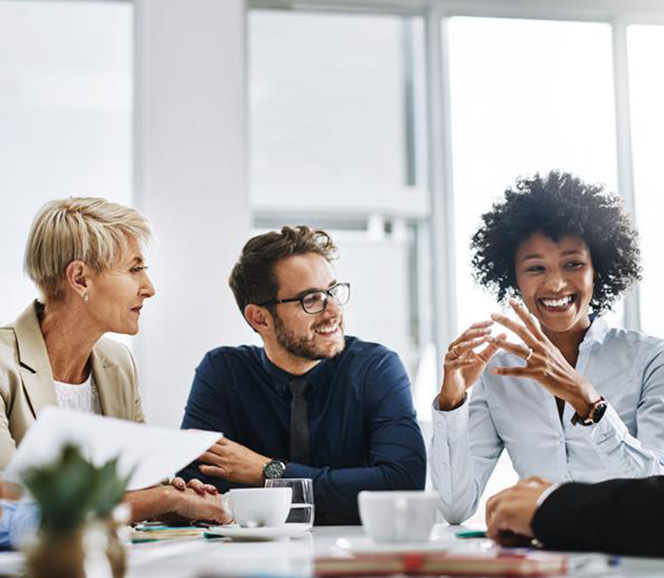 Three colleagues smiling and discussing around a conference table with coffee cups and documents.