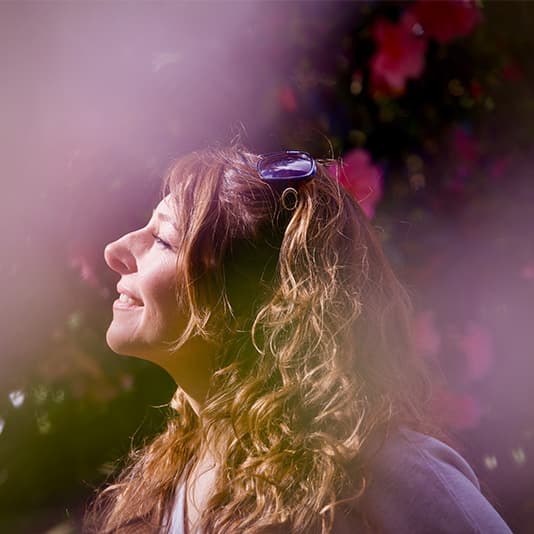 Woman with curly hair and sunglasses on head, smiling with eyes closed, surrounded by soft pink and purple hues, against a blurred background.