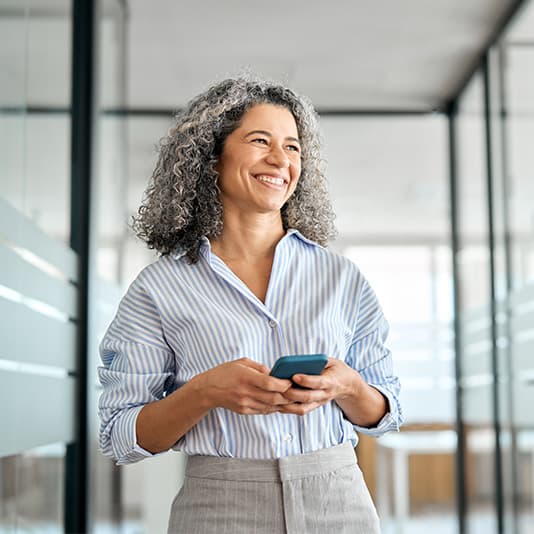 Smiling gray-haired woman in a striped shirt holding a smartphone in a bright, modern office corridor.