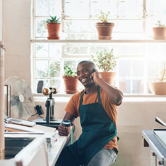 Man in a shop wearing an apron, smiling while talking on the phone, holding a cup. Behind him, potted plants sit on a sunny windowsill.