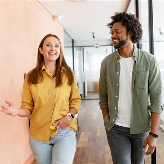Two people walking and talking in a modern office hallway, one wearing a yellow shirt, the other in a green shirt. Both appear cheerful.