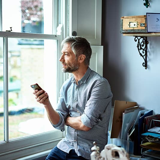 Man in a blue shirt holding a phone, looking thoughtfully out a window in a cozy room with shelves and boxes.