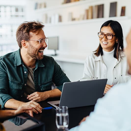 Two people in an office, smiling and talking at a table with a laptop open