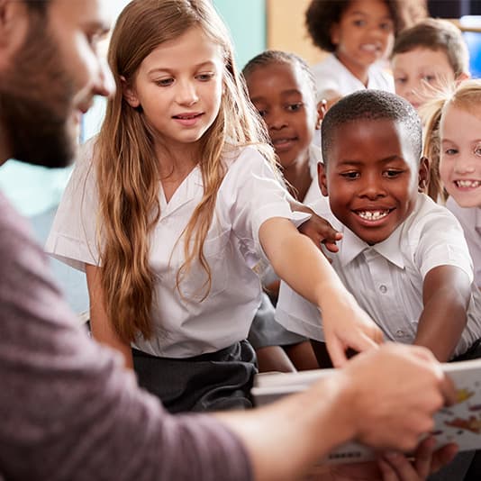 A diverse group of children in school uniforms eagerly gather around a teacher, pointing and smiling at a book he's holding.