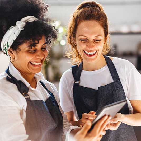 Two women in aprons smiling and looking at a tablet together in a kitchen or café setting.