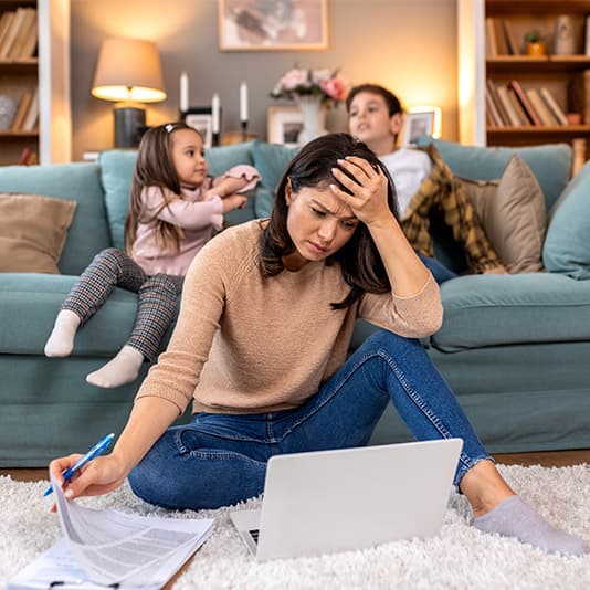 A stressed woman sits on the floor with a laptop, surrounded by two playful children on a sofa in a cozy living room.