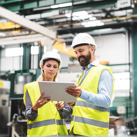 Two factory workers in safety vests and helmets review a document on a tablet in an industrial setting.