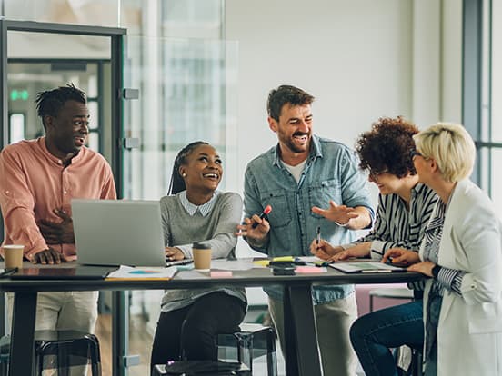 A diverse group of five people around a table, laughing and engaging in discussion in a bright office space.