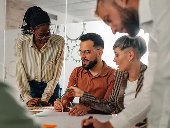 A diverse group of people collaborating at a table, discussing and pointing at documents in a modern office setting.
