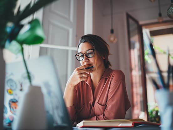 Woman in glasses, wearing a pink shirt, sits at a desk thoughtfully looking at a laptop, with a pen in hand. Open door and plants in background.