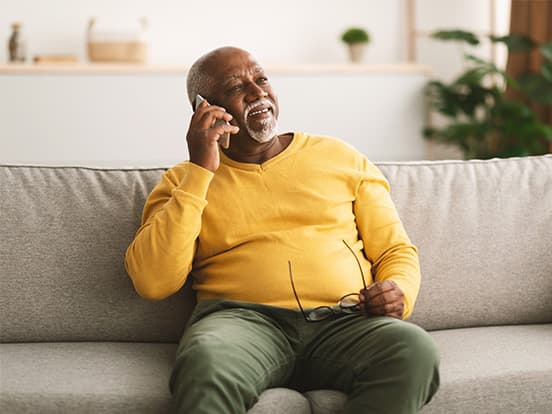 Smiling older man in a yellow sweater sits on a sofa, holding glasses and talking on a smartphone in a bright living room.