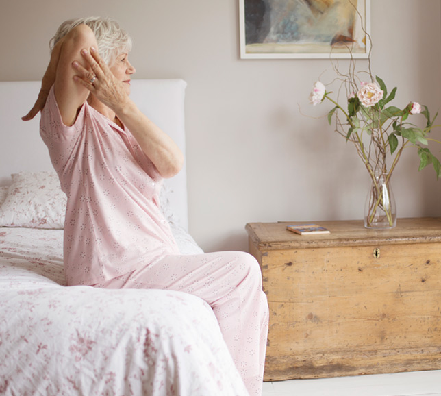 Elderly woman sat on the edge of her bed and stretching her arms