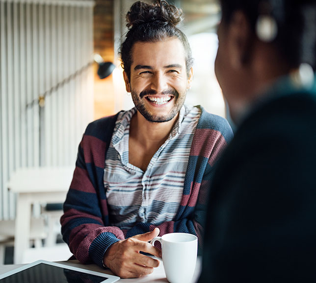 Man holding a mug and smiling at a friend across a table