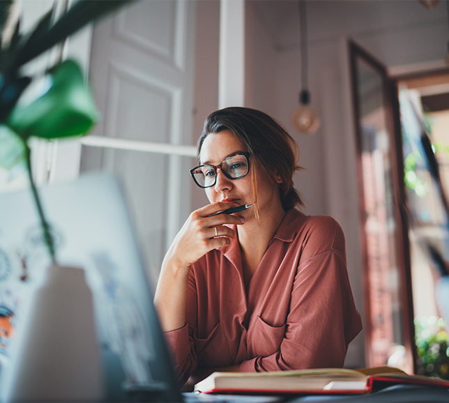 A woman in glasses, wearing a pink shirt, sits at a desk with a laptop, pen in hand, appearing thoughtful