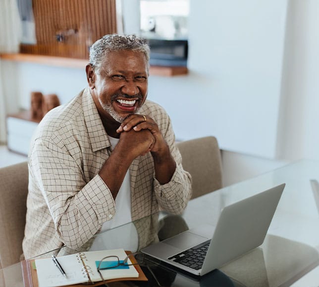Man sat at table with laptop and notepad, and smiling